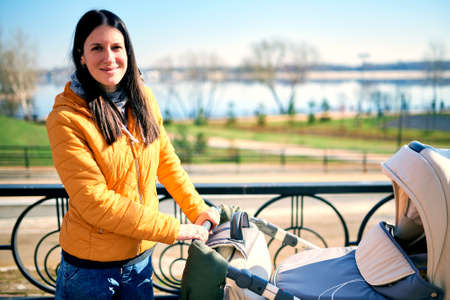 A happy young mother keeps a stroller with a child on the street to the park and looks into the camera. outdoor leisure. Sunny weather and beautiful scenery. Healthy lifestyleの写真素材