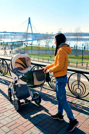 A happy young mother rolls a stroller with a child on the street to the park. leisure in nature. Sunny weather and beautiful scenery. Healthy lifestyleの写真素材
