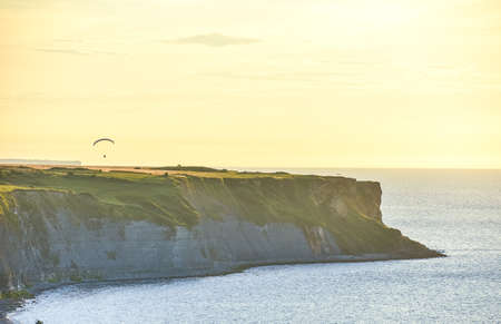 Rocky cliffs covered with green grass at sunset. A man practicing paragliding over them. Normandy,...