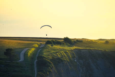 Rocky cliffs covered with green grass at sunset. A man practicing paragliding over them. Normandy, France.の写真素材