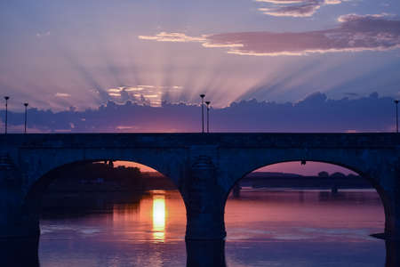 Stunning sunset with light coming through the clouds and a colorful sky. In the foreground a bridge of a city in backlighting.の写真素材