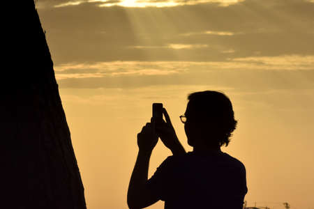 Backlit silhouette of an unrecognizable boy with long hair and glasses, taking a photo with his mobile phone. Lisbon, Portugal. Europe.の写真素材