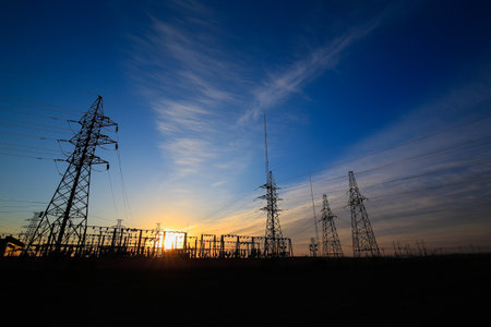 Many high-voltage power towers, silhouetted against the setting sunの写真素材