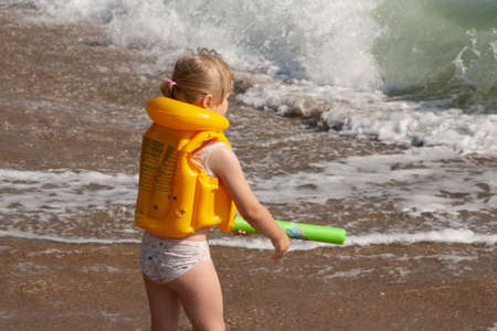 Young girl with yellow life jacket. On the shore with the waves.