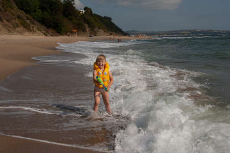 Young girl with yellow life jacket with water pistol. On the shore with the waves.の写真素材