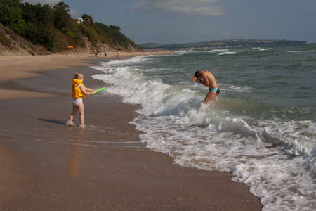 Two children a boy and a girl, a brother and a sister are playing with a water gun on the seashore in the water. Girl in a life-saving yellow vest. Bulgaria.の写真素材