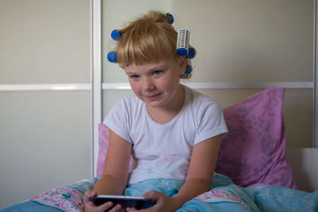 Sleepy girl with curlers in bed in the morning looks at the phoneの写真素材