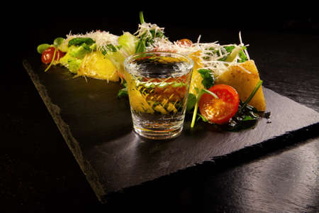 Cheese sliced with herbs and tomatoes with a glass of vodka on a dark backgroundの写真素材