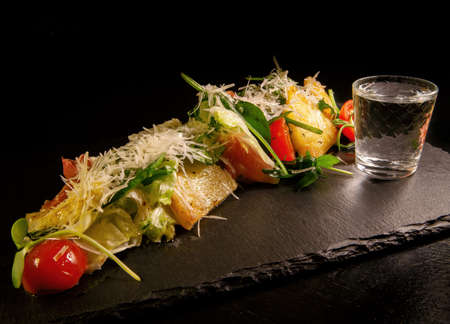 Cheese sliced with herbs and tomatoes with a glass of vodka on a dark backgroundの写真素材