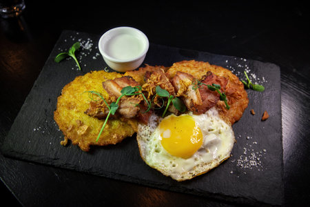 Restaurant serving snacks on the board - potato pancakes with fried meat and egg. A bowl with sauce and a glass of vodka in the background.の写真素材