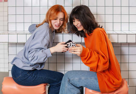 Two excited young girls using mobile phones while sitting at the cafe and pointing finger