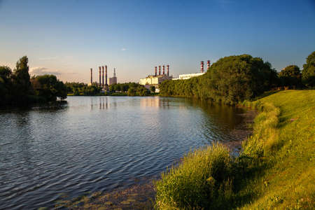 The landscape of a city park with a river and green trees, industrial production pipes in the...