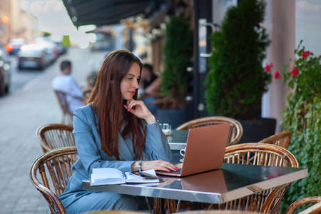 Close up portrait of smiling stylish european woman in white jacket sitting on street cafe and working on laptop.の写真素材