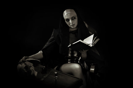 An evil cursed nun with a notebook and a cross in her hands looks at the camera on a black background. Halloween. Horrors.の写真素材