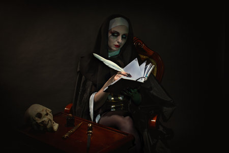 A nun, in typical black and white clothes, writes in a notebook with a pen, sitting in an armchairの写真素材