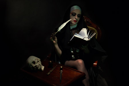An emotional woman dressed for Halloween as a nun sits on a black background in an armchair with a notebook with a penの写真素材