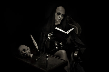 An emotional woman dressed for Halloween as a nun sits on a black background in an armchair with a notebook with a pen.の写真素材
