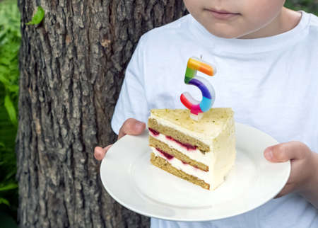 A boy in a white T-shirt holds a piece of cake with a candle on a plate against the background of...