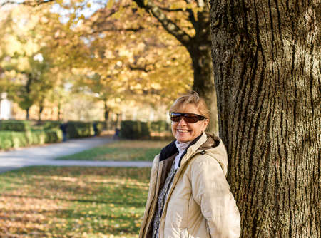 Mature, smiling woman in autumn park at treeの写真素材