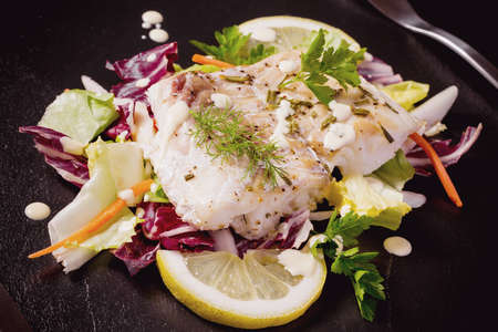Grilled cod fillet with salad on black slate plate, top view