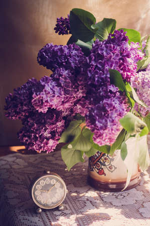 Lilac flowers in a ceramic pot with vintage tiny alarm clock on white lace surfaceの写真素材