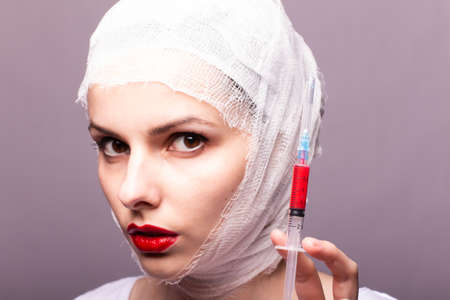 beautiful young girl, lips painted up with red lipstick, head bandaged with a bandage, in the hands of a syringe with a red liquidの写真素材