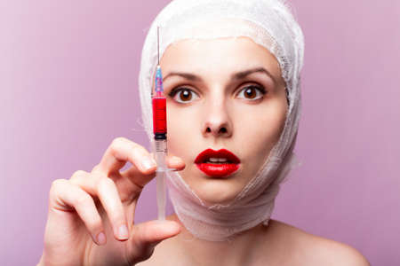 beautiful young girl, lips painted up with red lipstick, head bandaged with a bandage, in the hands of a syringe with a red liquid, pink backgroundの写真素材