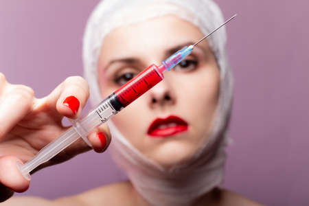 beautiful young girl, lips painted up with red lipstick, head bandaged with a bandage, in the hands of a syringe with a red liquid, pink backgroundの写真素材