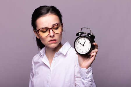 woman manager in a white shirt and glasses holds a clock, morning in the office, gray studio backgroundの写真素材