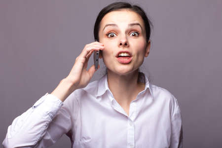 cute businesswoman in a white shirt is emotionally talking on the phone, gray backgroundの写真素材