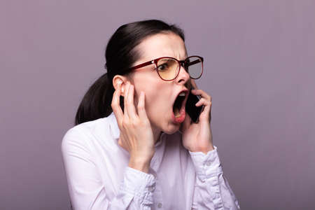 young woman office manager in glasses and a white shirt speaks on the phone, angry, gray backgroundの写真素材