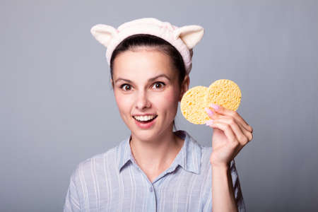 Image of young woman with fresh skin and young outlook, covering eye with cosmetic sponge, wears...