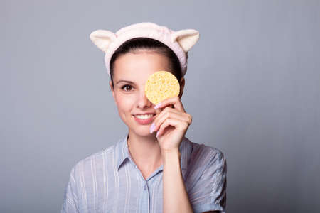 Image of young woman with fresh skin and young outlook, covering eye with cosmetic sponge, wears gray casual shirt, takes care of beauty, isolated over white backgroundの写真素材