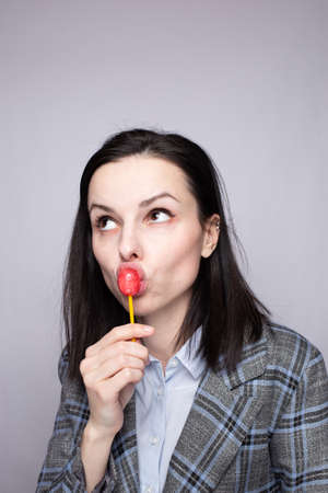 Woman office worker in gray plaid suit eating lollipop, gray studio background