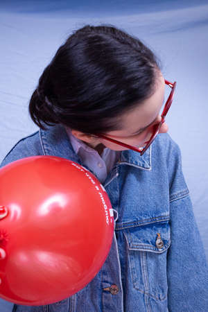 woman with red lips, in glasses and with an inflatable ball in her hands, dressed in a blue shirt, denim jacket on a blue backgroundの写真素材