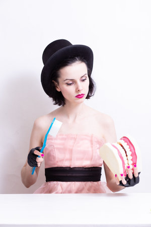 A woman in a dress and hat sits at a table and holds a mock-up of her teeth. high quality photo