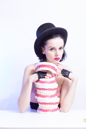 A woman in a dress and hat sits at a table and holds a mock-up of her teeth. high quality photo