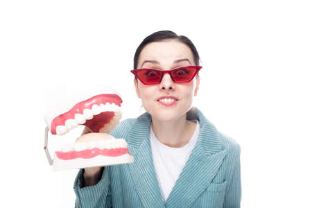 enthusiastic woman in red glasses and corduroy jacket holds a dental jaw in her hand, white background. high quality photoの写真素材