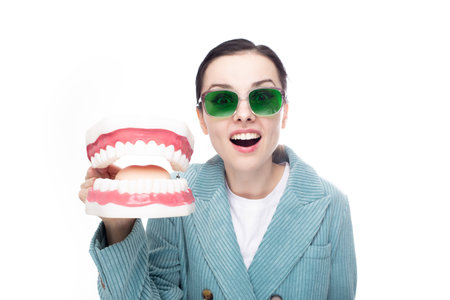 cute surprised woman in green glasses and a jacket holds a large dental jaw in her hand. high...
