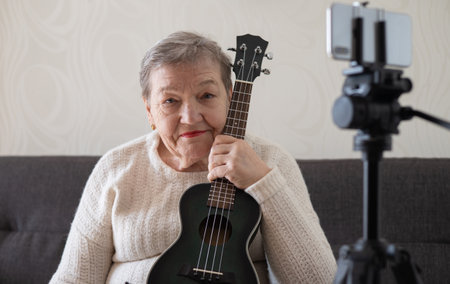 Elderly woman, grandmother with a ukulele in her hands sits on the couch next to smartphone on tripod and thinks about something. She had video chat with relatives. Adaptationの写真素材