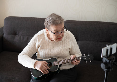 Elderly woman 70+ sits on the couch at home and plays the ukulele Adaptation of pensioners in the modern world. New knowledge and skills, hobby.の写真素材