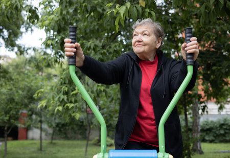Smiling elderly woman is doing exercises on simulator, trainer ellipse in public park. Active life of pensioners. Adaptation in the modern world. Prevention of mental illness.の写真素材