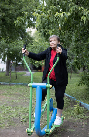 Smiling elderly woman is doing exercises on simulator, trainer ellipse in public park. Active life of pensioners. Adaptation in the modern world. Prevention of mental illness.の写真素材