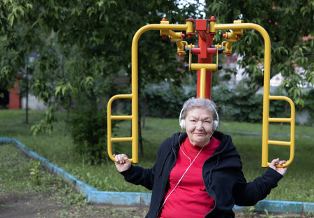 Smiling elderly woman in headphones is doing exercises on simulator, trainer outdoors in the yard....