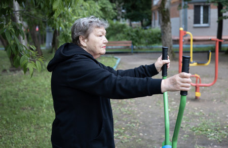 Smiling elderly woman is doing exercises on simulator, trainer ellipse outdoors in the yard. Active...
