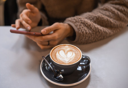 Girl in fluffy sweater holds a smartphone in hands and writes message next to a cup of coffee with heart shaped latte art foam. Close up cup of coffee with cream in coffee shop.の写真素材