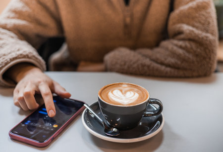 Girl in fluffy sweater holds a smartphone in hands and writes message next to a cup of coffee with heart shaped latte art foam. Close up cup of coffee with cream in coffee shop.の写真素材