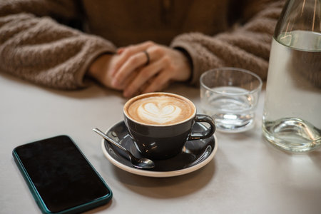 Girl in cafe waits for call from loved one over cup of coffee with heart shaped latte art foam....