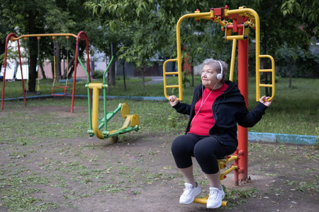 Smiling elderly woman in headphones is doing exercises on simulator, trainer outdoors in the park. Active life of pensioners. Adaptation of pensioners in the modern worldの写真素材