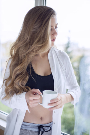beautiful sports girl near window with mug in hands. morning in a shirtの写真素材